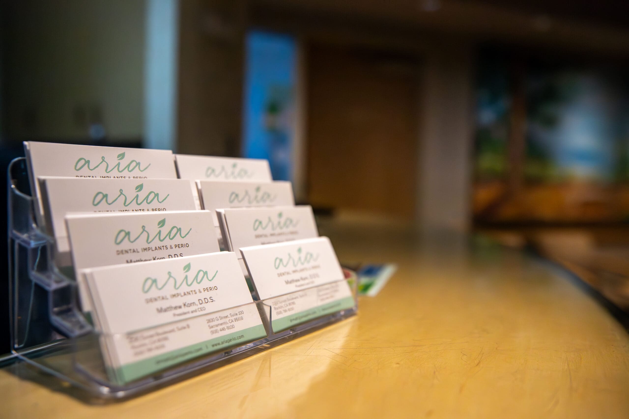 Acrylic holder displaying stacked “arria” dental practice business cards on a wooden counter.